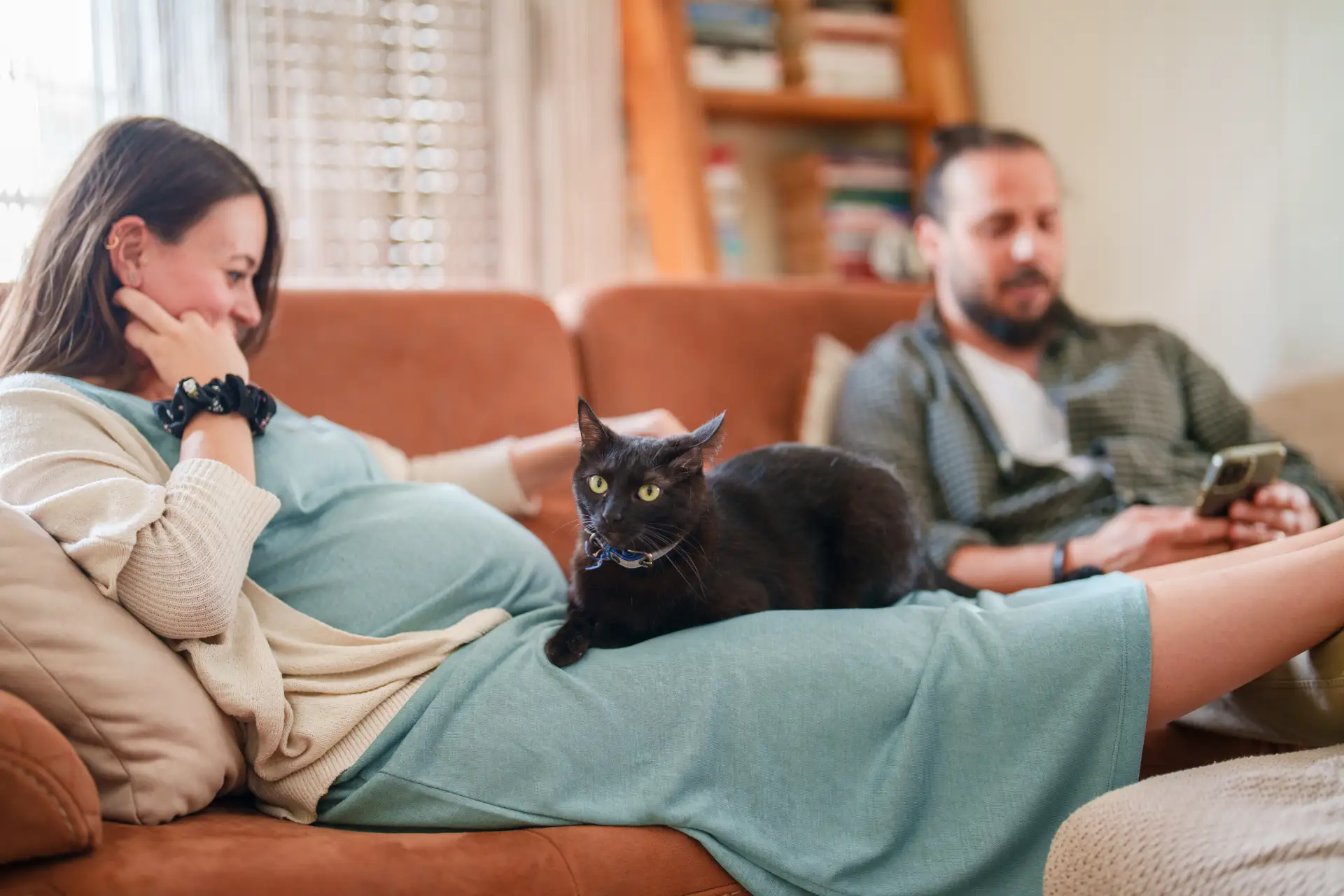 A pregnant woman in a green dress lounging on a rust colored couch next to her husband with a black short haired cat in her lap.